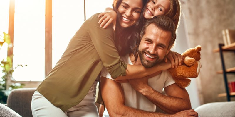 Happy dad and mom with their cute daughter and teddy bear hug and have fun sitting on the sofa in the living room at home.