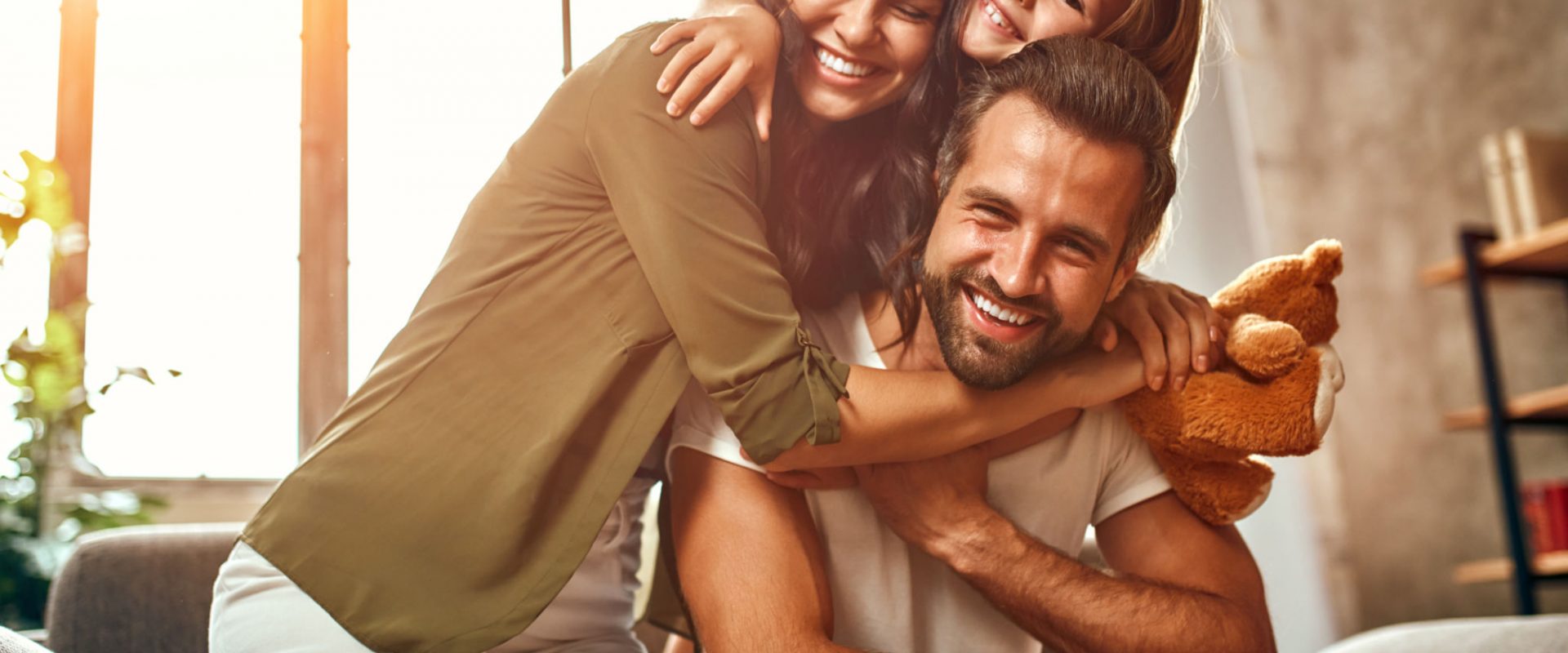 Happy dad and mom with their cute daughter and teddy bear hug and have fun sitting on the sofa in the living room at home.